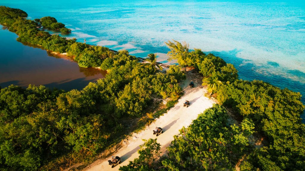 ATV adventure along a mangrove-lined coastal trail in Grand Cayman, with shallow coral flats and turquoise Caribbean waters beyond.